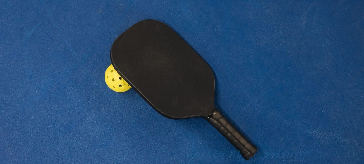 A black beginner pickleball paddle resting on a yellow ball on a blue court surface.