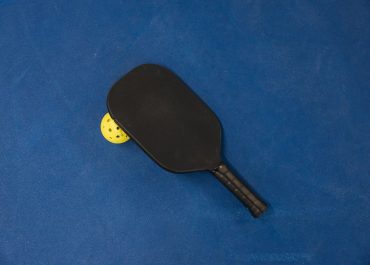 A black beginner pickleball paddle resting on a yellow ball on a blue court surface.