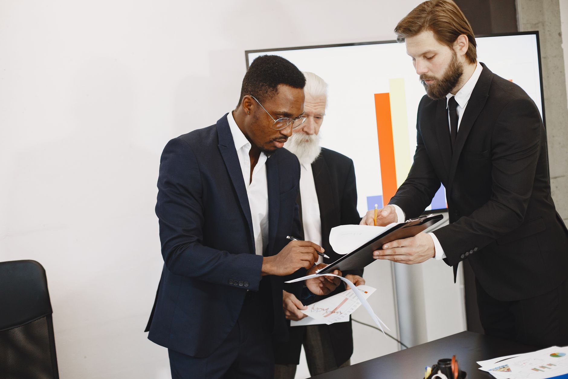 Three businessmen engaged in document signing in a modern office setting. — Brett Hartvigson: How Working With A Financial Ad