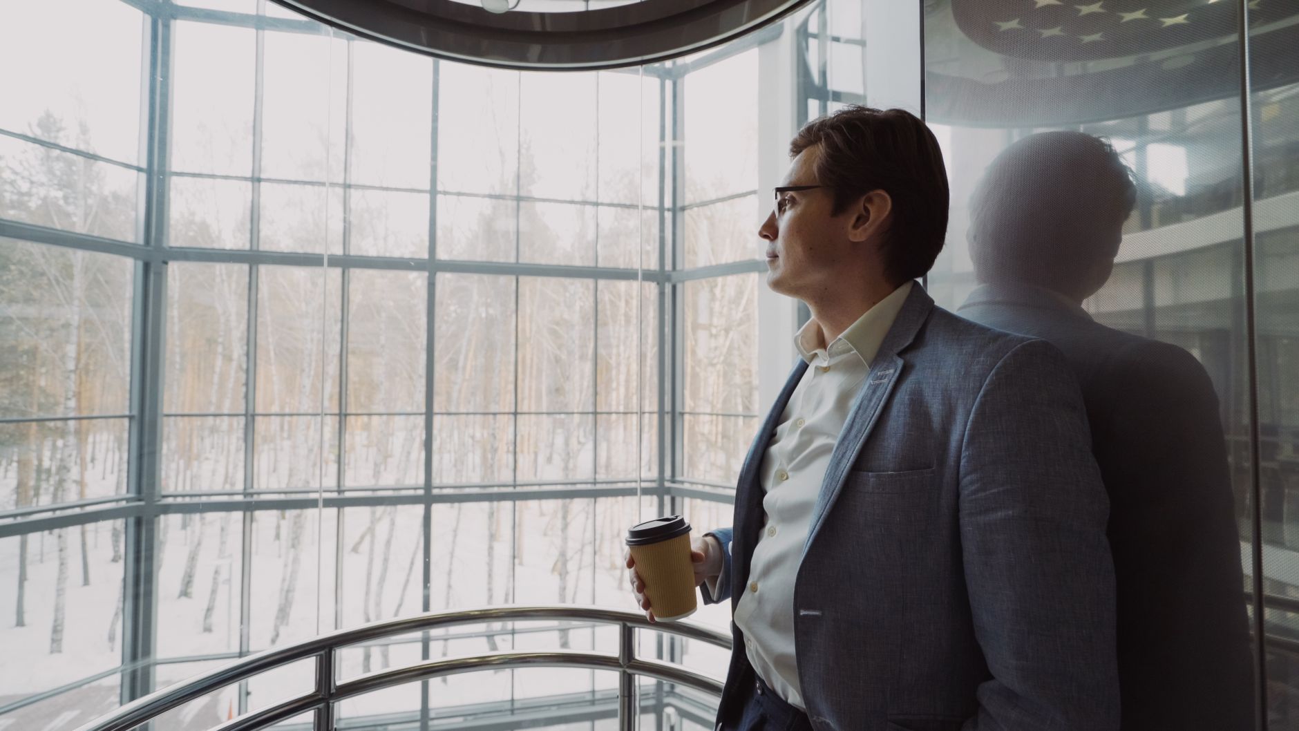 Businessman in blue suit with coffee cup looking out a large window, reflecting on a winter landscape. — Discover Comfortable