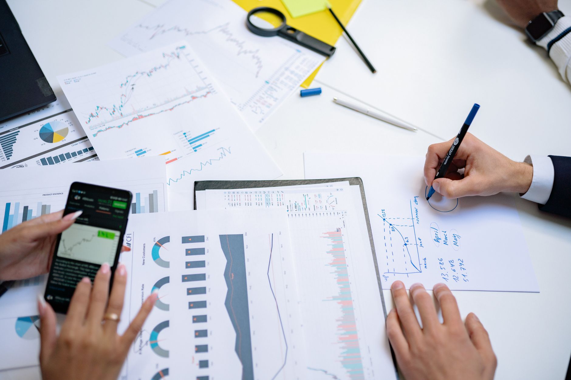 Colleagues collaborating on financial charts and graphs using mobile and paper documents in the office. — Dr Wade Banker - Tr