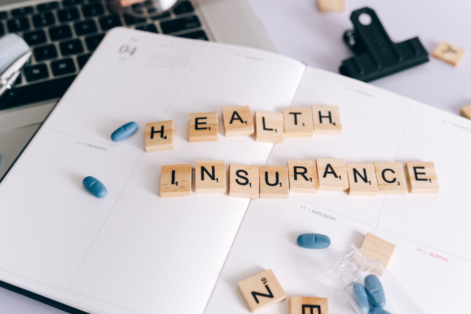 Scrabble tiles spelling 'Health Insurance' on planner with pills and laptop, symbolizing healthcare planning. — Transformativ