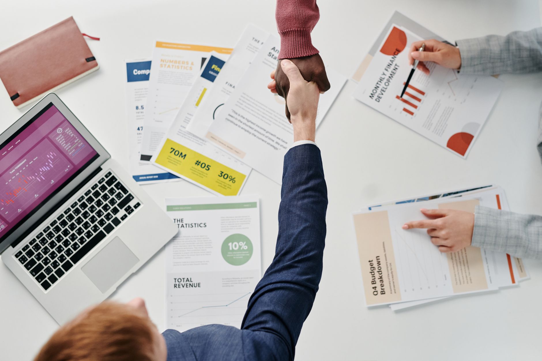 Business professionals engaging in a handshake over a desk filled with financial documents and a laptop. — Unraveling The Eni